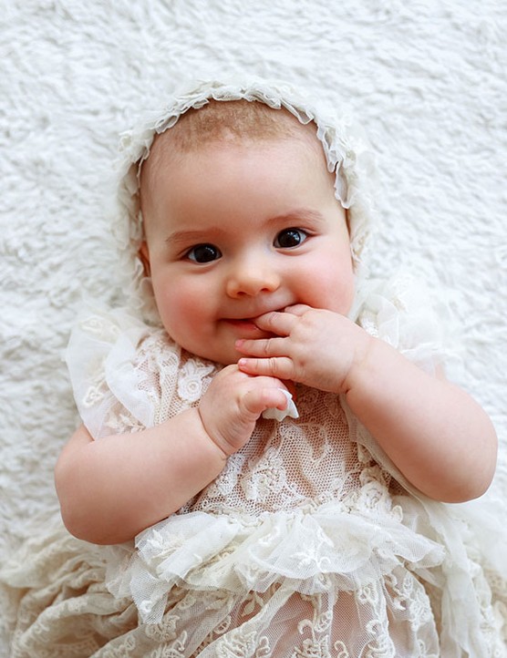 little child lies on the bed and smiles baby girl lying down wearing an old fashioned dress and bonnet