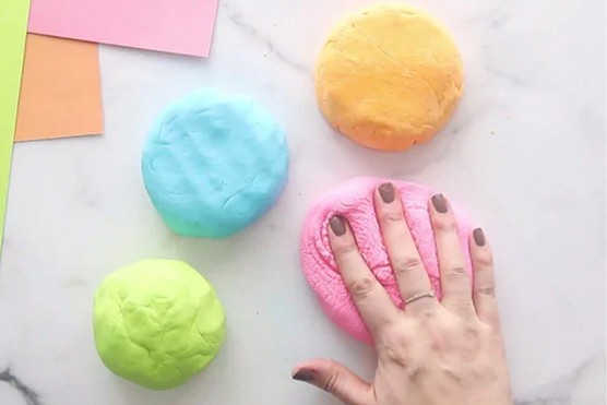 4 pieces of cloud dough in yellow, blue, green and pink with a hand pressing into the pink dough