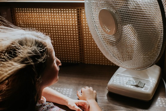 A little girl sits at a table indoors in front of a desk fan blowing in her face.