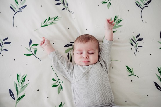 Baby girl stretching her arms with her eyes closed, lying on a patterned sheet.