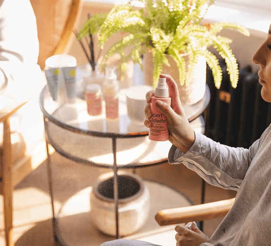 A woman sitting in a chair, holding a bottle of My Expert Midwife's Spritz for Bits